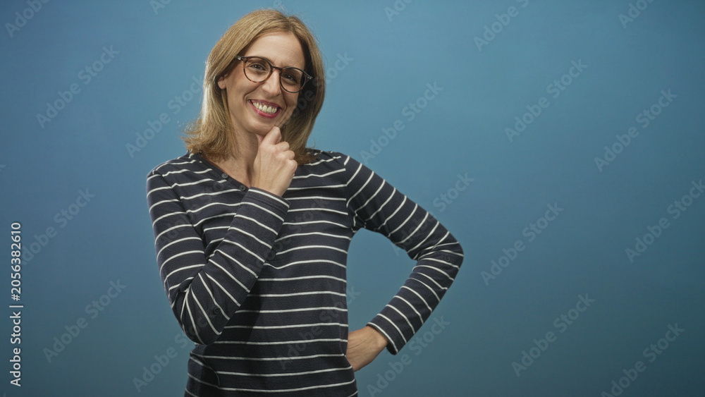 Fototapeta premium Woman wearing glasses and striped top, blonde, finger to chin and hand on hip while smiling in studio; friendly confidence.