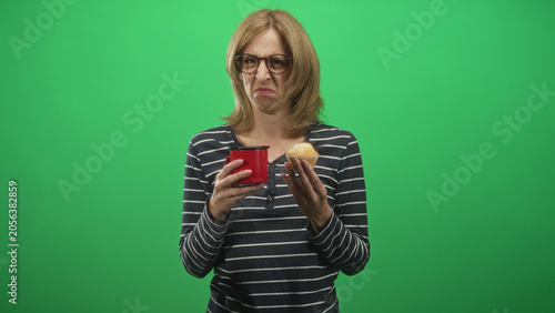 Fotografía Woman holding red mug and muffin grimacing in studio with green screen, wrinkled nose showing distaste; disgust morning routine refusal