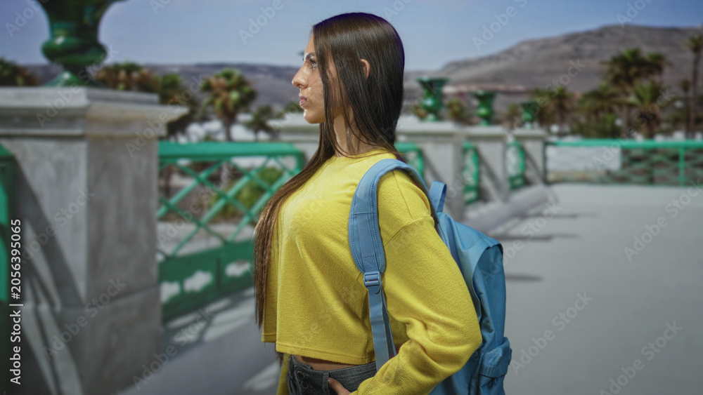 Obraz premium Young woman student in yellow sweater with blue backpack stands in profile, hand in pocket, on a street promenade by the sea; contemplation.
