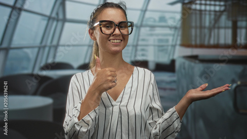 Obraz na plátne Young woman wearing glasses and striped blouse points finger to open palm in building restaurant interior while smiling and offering gesture; confidence