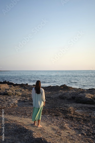 Distant view of a woman standing on a rocky beach in Paphos and looking at the sea at sunset