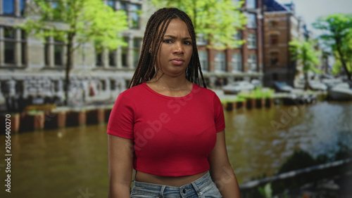 Fotografía Woman scowls at camera on canal street in amsterdam wearing red shirt and jeans, standing by water and bridge; urban annoyance