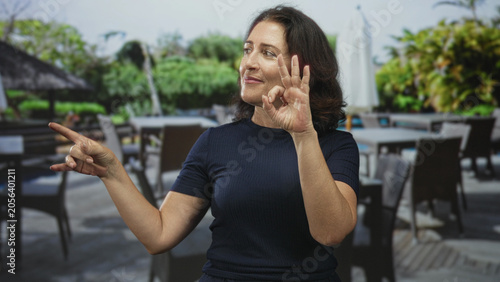 Foto Woman showing ok sign with right hand and pointing finger left toward patio tables and chairs at outdoor restaurant building; confidence connection