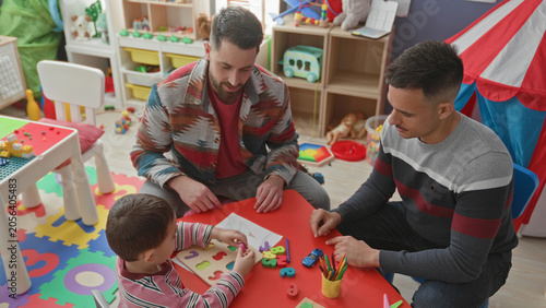 Photos Young boy places wooden puzzle pieces while two men guide with hands at a colorful play table in a building; warmth bonding