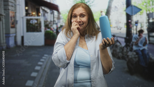 Φωτογραφία Woman holds blue soap bottle in right hand and touches chin with left, hesitant facial expression in a city street outside a cafe and parked bicycle; uncertainty decision