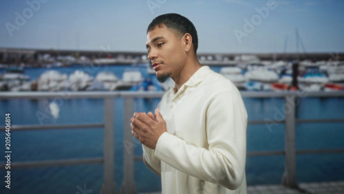 Foto Young hispanic man with hands clasped, rubbing palms near marina railing, facing moored boats while speaking; contemplation