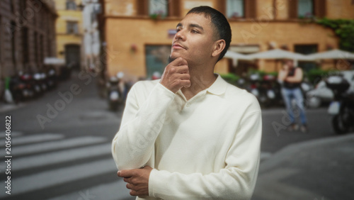 Εκτύπωση καμβά Man touching chin with hand gesture on an urban street by a cafe building and parked scooters; thoughtful reflection