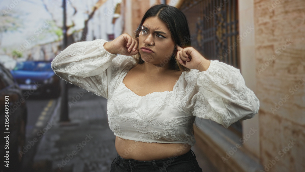 Fototapeta premium Woman plugging ears with fingers and pouting, white embroidered crop top and black jeans visible on a narrow street with parked car and iron gate; annoyance discomfort.