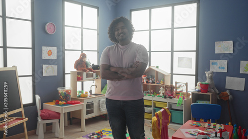 Foto Man arms crossed standing amid toys and cubbies in a kindergarten building, young teacher smiling at camera; nurturing calm learning