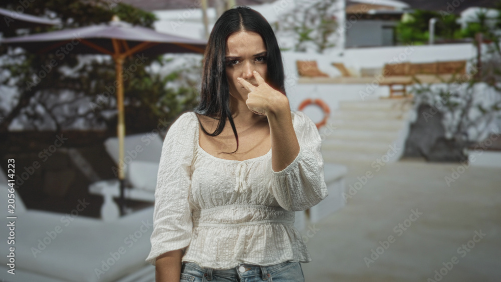 Fototapeta premium Young hispanic woman with crossed arms and frowning face near a resort pool building; frustration defiance.