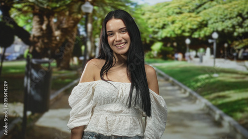 Fotografie Young woman smiling with bare shoulder visible, posing on a sunlit park pathway under trees; youthful joy