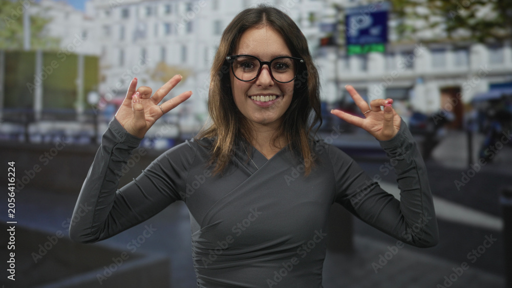 Fototapeta premium Woman showing double peace signs with fingers and hands on a busy city street with buildings and parked cars; joy youthful playfulness.