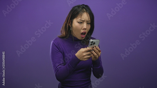 Tablou pe pânză Woman holding smartphone with both hands, startled frown, arms extended and pointing phone forward against a purple studio wall; surprise concern