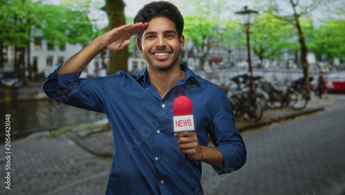 Billede på lærred Man holding red microphone labeled news, smiling and addressing camera on street by canal with bicycles; news reporting confident