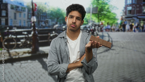 Obraz na plátně Young hispanic man holding a chocolate bar to his face with crossed arms on a street canal bridge, looking pensive and unsure; disappointment longing