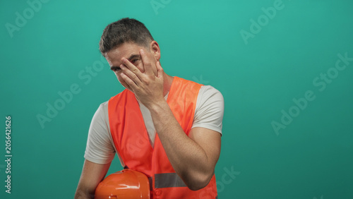 Photography Man engineer in reflective orange vest holding orange hardhat and covering face with hand in teal studio; embarrassment