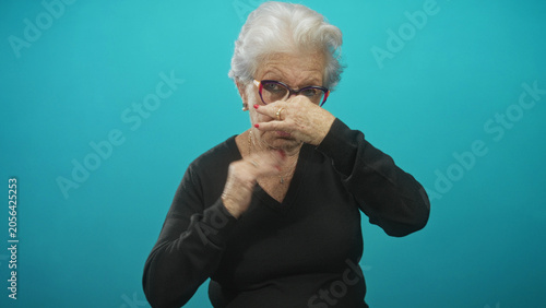 Φωτογραφία Elderly woman in black sweater pinches her nose with hand and closes eyes in turquoise studio beside glasses; disgust aversion