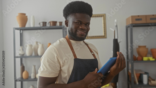 Fotografija Man artisan in apron holds tablet with fingers on screen in pottery studio among shelves of ceramic vases and clay pots; craft pride focus
