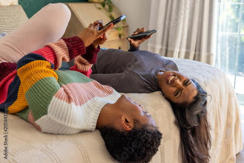 Two smartphones are resting on textured bedspread near wooden dresser and sheer curtains