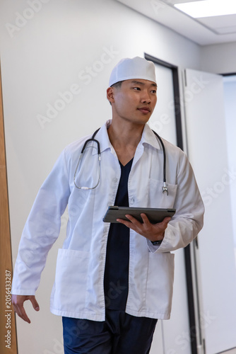 Asian male physician walking in clinic hallway holding tablet and stethoscope, wearing lab coat
