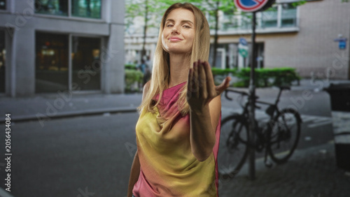 Canvas-taulu Woman beckons with hand, palm up and slight smile on a city street by parked bicycles and storefront windows; playful invitation