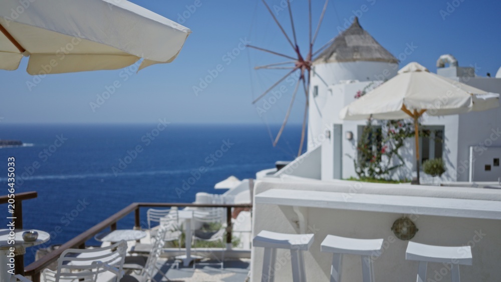 Fototapeta premium Seaside terrace with white chairs, umbrellas and distant windmill in soft defocused bokeh outdoor background; backdrop copyspace calm.