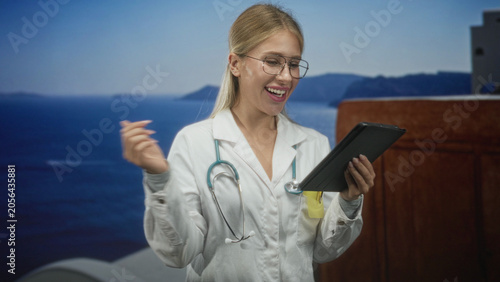 Fotografia Woman doctor in white coat taps tablet with finger in studio, stethoscope visible and smiling; telemedicine care cheerful