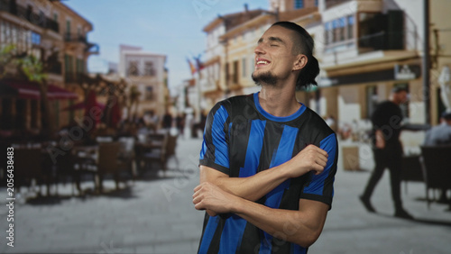 Canvastavla Man in blue and black soccer jersey hugs chest and smiles while gesturing on a sunny pedestrian street; joy celebration