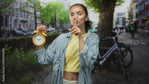 Fotografija Woman holding yellow alarm clock, index finger to lips for silence on a street beside a canal and parked bicycle near city buildings; urgent secrecy