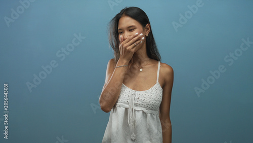 Fotografie Young brunette woman covers mouth with hand while smiling, wearing white crochet dress and bracelet in studio; shyness tenderness