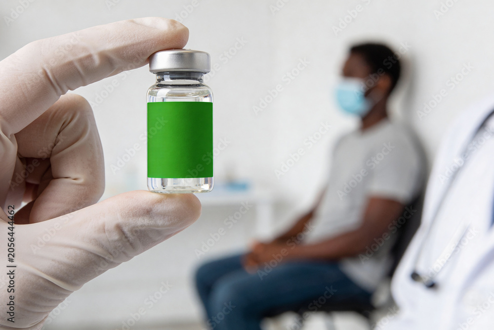 Obraz premium Doctor holding blank vaccine ampoule in front of African American patient wearing medical mask. Development of remedy against Ebola and measles. Healthcare and immunization concept.