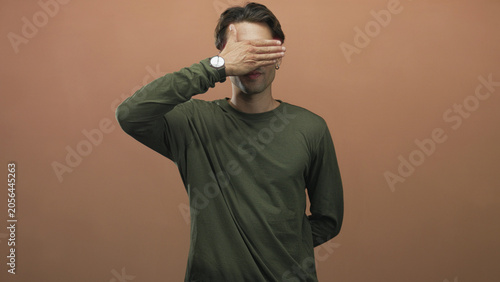 Принти на полотні Man covers eyes with hand showing wristwatch and earring, smiling in olive green longsleeve shirt in studio; playful shyness