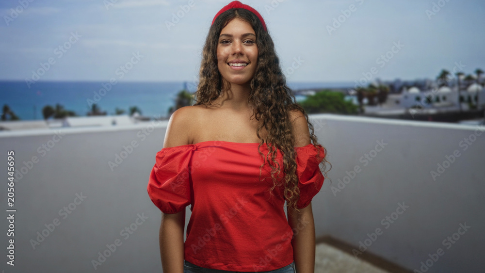 Fototapeta premium Teenage hispanic woman with bare shoulders in a red off shoulder top smiling and looking up on a rooftop building terrace by the sea; carefree joy.