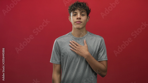 Foto Teenage man places hand on chest with eyes closed and calm expression against red studio backdrop; sincerity gratitude