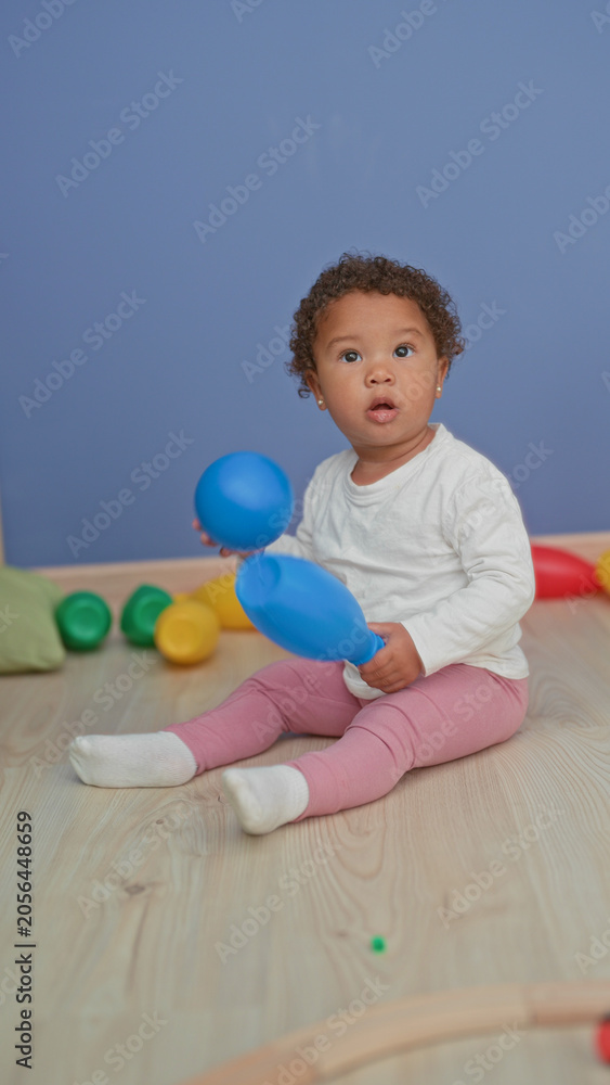Naklejka premium Little girl wearing white shirt and pink leggings puts a blue ball to her mouth while sitting on a wooden floor surrounded by colorful toy pins in a studio; curiosity discovery play.