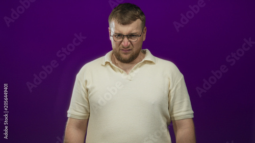 Fotografie Man with glasses grimacing and squinting in studio against purple backdrop wearing cream polo shirt, hands down by sides; disgust discomfort