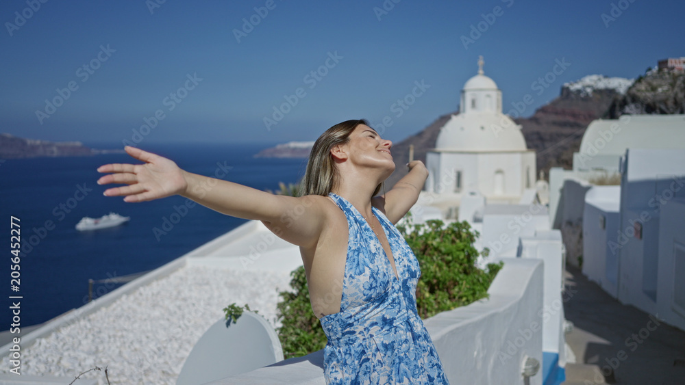 Obraz premium Woman smiling with arms outstretched on a white church building terrace in santorini overlooking blue sea and island vistas; serenity escape.
