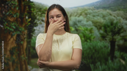 Tableau sur toile Woman in pale yellow t shirt with hand covering mouth and arm crossed at elbow in forest among lush green foliage; shyness warmth