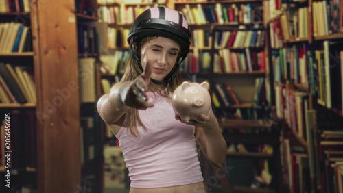 Fotografie Woman points finger to camera holding piggy bank wearing helmet and crop top in library; saving confidence