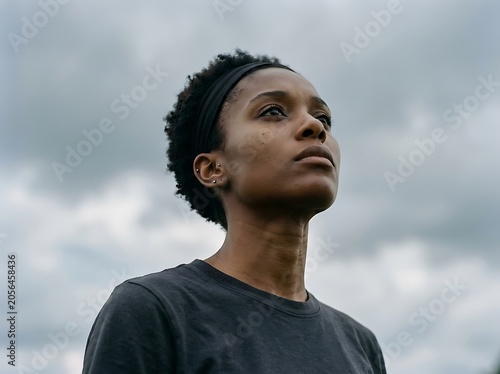 Close-up of a determined young Black woman looking upwards with hope and confidence against a cloudy sky, exuding strength and resilience