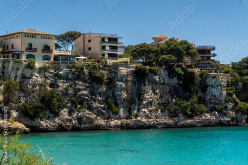 Beautiful view of a turquoise sea bay with houses built on rocky cliffs surrounded by green trees under a clear blue sky on a sunny summer day.