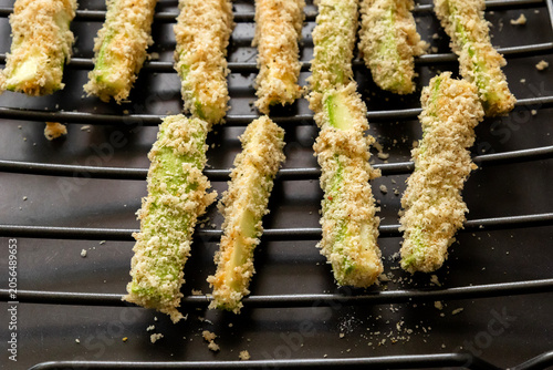 Close-up of raw zucchini fries coated in parmesan panko breadcrumbs and spices, neatly arranged on a wire rack ready for baking