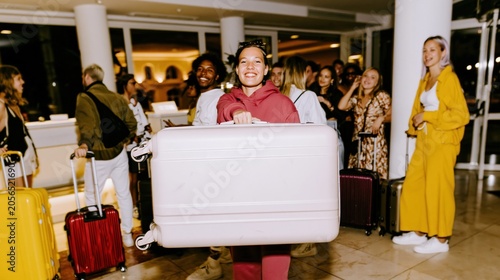 Smiling young woman holding a large hard-shell suitcase while arriving at a luminous hotel lobby with a group of friends for a summer vacation