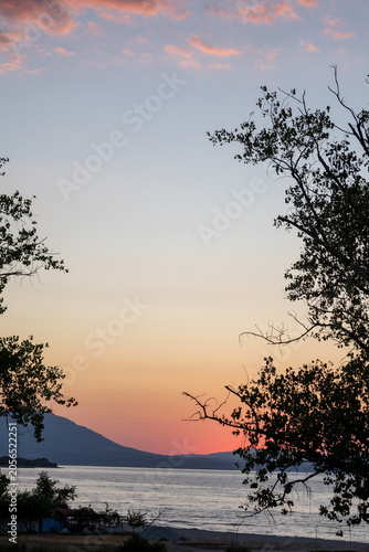 Sunset sea view framed by trees.Sunset over calm sea with a mountain silhouette framed by trees and soft pink clouds in a peaceful coastal landscape