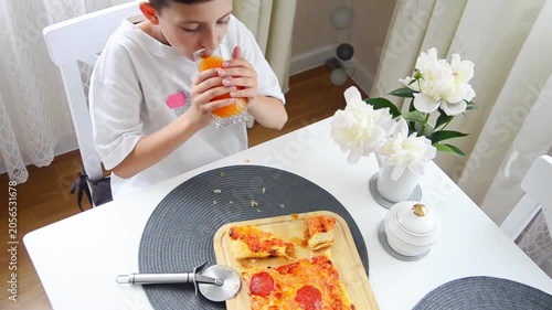 Young boy drinks a glass of orange juice while sitting at the kitchen table during lunch. Cozy family mealtime scene with homemade pizza, healthy beverage and bright natural daylight.