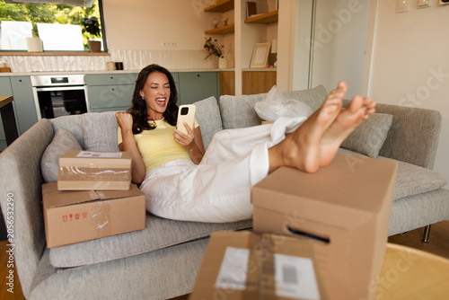 Happy woman relaxing among many cardboard boxes, checking smartphone and raising victory sign after receiving online orders at home