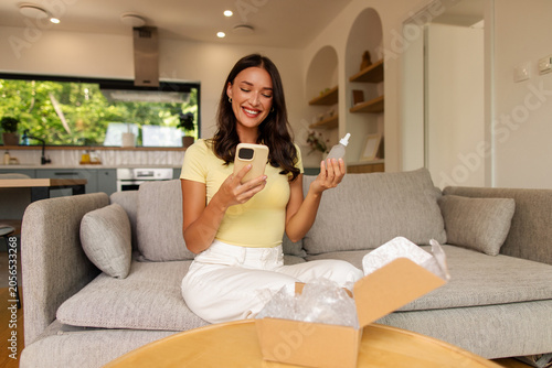 Happy woman looking at smartphone with smile and holding skincare serum while opening delivery box filled with cosmetic products on sofa