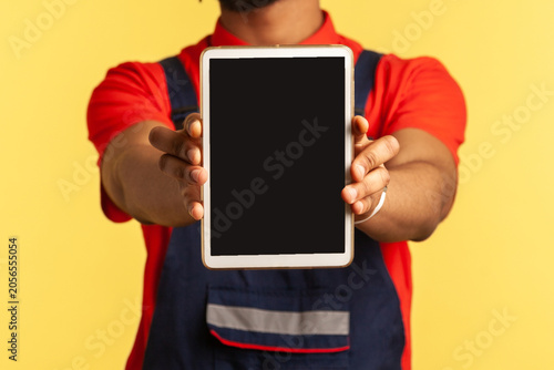 Happy handyman wearing blue overalls holding out showing tablet with blank screen with copy space for promotion, advertising industry service. Indoor studio shot isolated on yellow background.