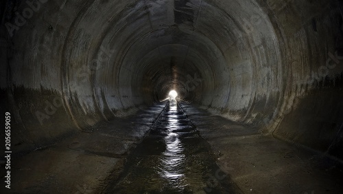 Inside a dark urban drainage culvert, wet walls leading to distant light.