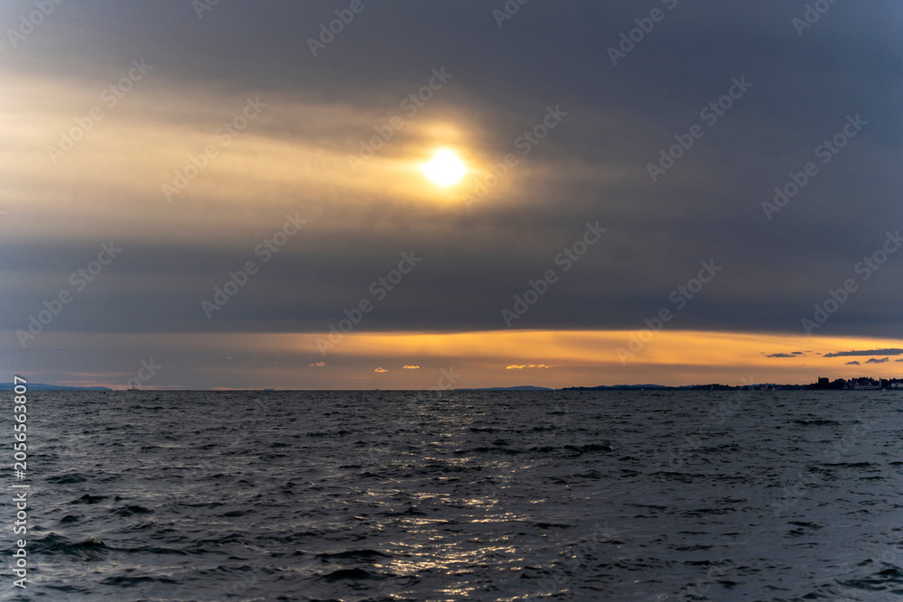 Obraz premium A dramatic sunset over Lake Constance viewed from Bregenz, featuring dark storm clouds parting to reveal a golden glowing sun and light reflecting on the deep blue water surface